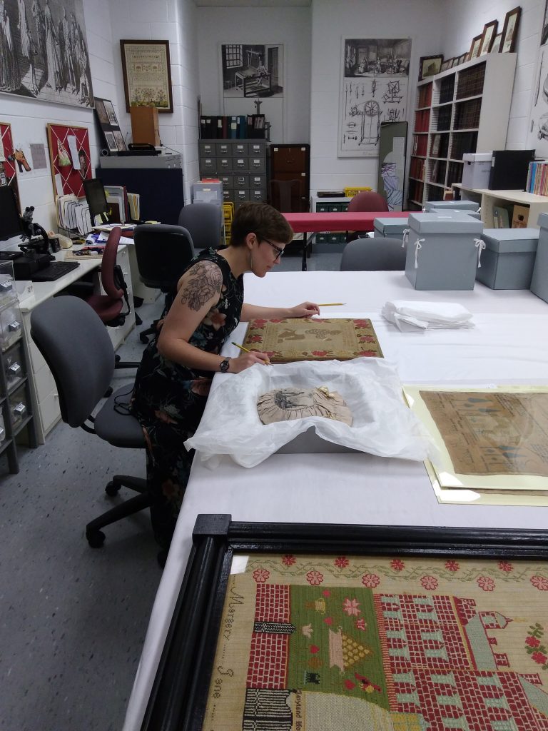 Figure 2: Mariah Gruner at Colonial Willamsburg examining an abolitionist sampler of unknown origin and date (likely ca. 1840-55) and a workbag made by the Female Society for Birmingham, West Bromwich, Wednesbury, Walsall, and their Respective Neighbourhoods for the Relief of British Negro Slaves in 1827.