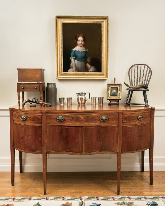 The Frankfort sideboard dates to 1790. Primary wood is figured cherry with inlaid decoration with bell flower drops. Left to right: miniature sugar chest, flintlock pistol c. 1780, belonging to Colonel William Crabtree, stoneware by Isaac Thomas, coin silver pitcher by Asa Blanchard, coin silver beakers by Thomas Marsh and George McDannold, miniature on ivory by Matthew Harris Jouett, child’s Windsor chair from the estate of noted Kentucky Collector, Mrs. Eleanor Offutt, Kentucky powderhorn with applied antler tip from Robertson family. On the wall, “Nannie” by her uncle, Joseph Henry Bush.
