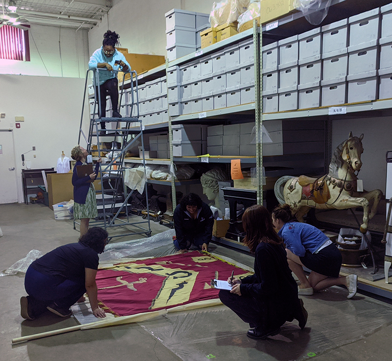 Cianni Williams (center), IDEAL intern at the Atwater Kent Collection at Drexel University, helps catalog a c. 1911 banner used in historical pageants created by and staged at the former John Wanamaker Department Store.