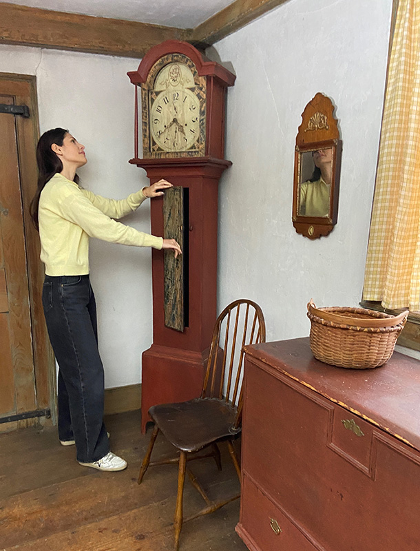 Figure 4. Livy Scott examines the painted surfaces of a clock in the Ashley House’s South Parlor. Silas Hoadley, Tall case clock, 1820–30, Plymouth, CT. Gesso, iron, ivory, paint, pewter, pine, tin. The Trustees of Reservations, AH.292. Image courtesy of The Trustees of Reservations.