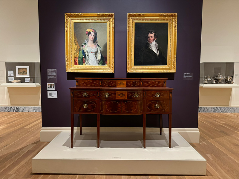 Figure 1. Installation at the Gibbes Museum of Art. Thomas Sully, Mr. and Mrs. Robert Gilmor, Jr., 1823. Oil on Canvas. Gibbes Museum of Art, Bequest of Lila Landson Jones, 1942.010.0003. Sideboard, 1790-1810, Charleston, SC. Mahogany and mahogany veneer; ash, poplar, and white pine. Gibbes Museum of Art, Gift of the Rivers Collection, 2023.003.0011.